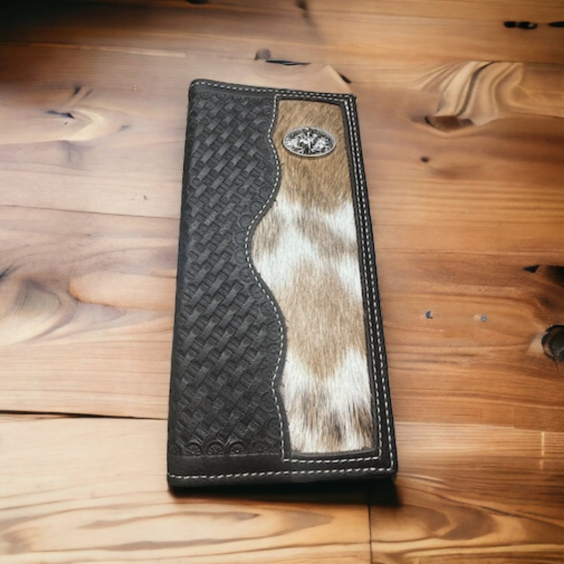 A Bull Rider Long Wallet W Cowhair Basketweave by Western Stakes, perfect as a cowboy accessory, lies on a wooden surface. The wallet features a woven black leather design on the left side, a strip of natural animal hide with fur running vertically down the middle, and a small metallic emblem in the top right corner.