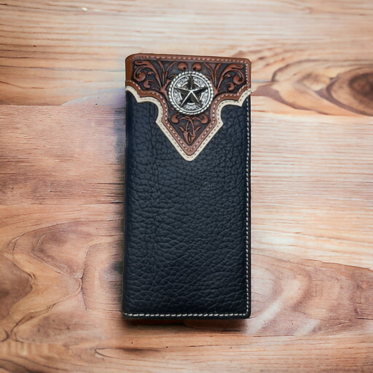 A Western Stakes Floral Long Wallet Black Star Concho with intricate brown detailing at the top, featuring a silver metal emblem with a star design. The wallet is placed on a wooden surface with visible grain patterns and has hints of floral design intricacies.