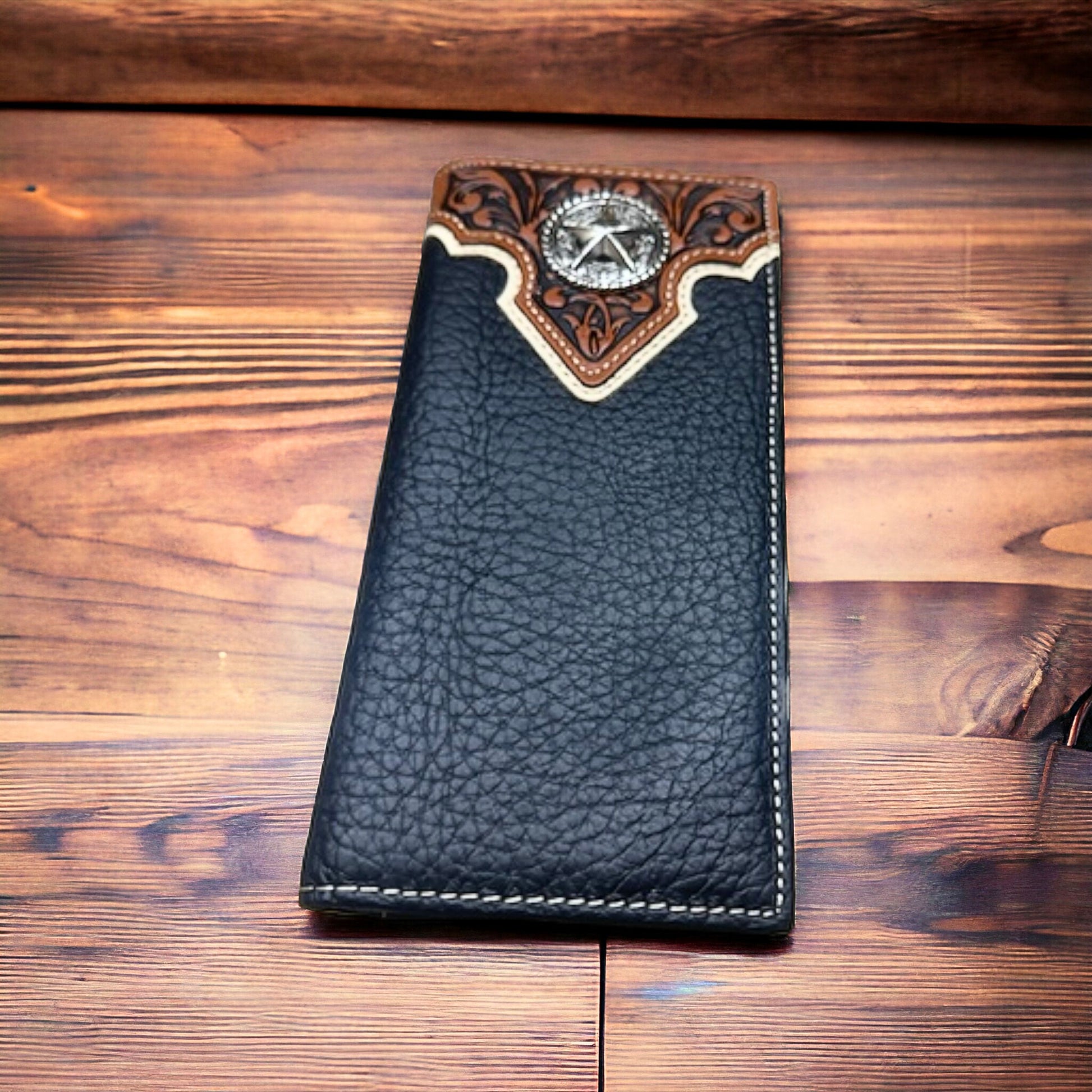 A Western Stakes Floral Long Wallet Black Star Concho with a decorative top flap featuring intricate brown and tan floral patterns and a silver star concho is placed on a wooden surface with a warm, rustic finish. The men's wallet has detailed stitching along the edges.