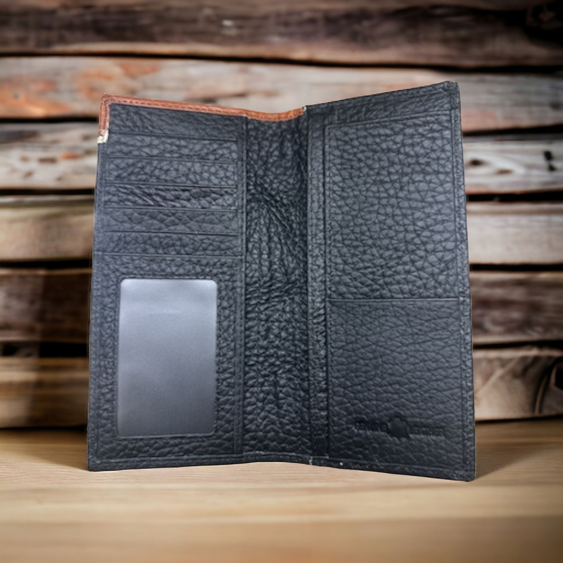 A Floral Long Wallet Black Star Concho by Western Stakes is shown open on a wooden surface with a rustic wooden background. The checkbook-style wallet features multiple card slots, an ID window, and pockets for cash and documents. The texture of the leather is prominently displayed.