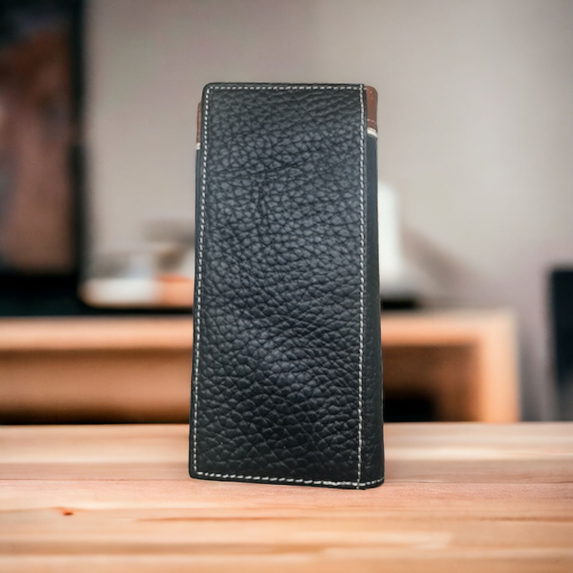 A rectangular black leather sleeve with visible white stitching, reminiscent of a mens wallet, stands upright on a wooden surface. The background appears slightly blurred, featuring a warm indoor setting with indistinct furniture. This item is the Floral Long Wallet Black Star Concho by Western Stakes.