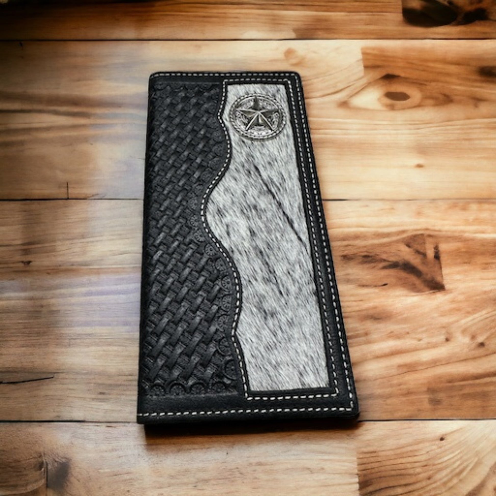 A Lone Star Long Wallet Black W Cowhair Basketweave from Western Stakes with intricate black woven leather on the left, gray fur texture on the right, and a silver star emblem at the top is placed on a polished wooden surface. This stylish cowboy accessory features white stitching along the edges.