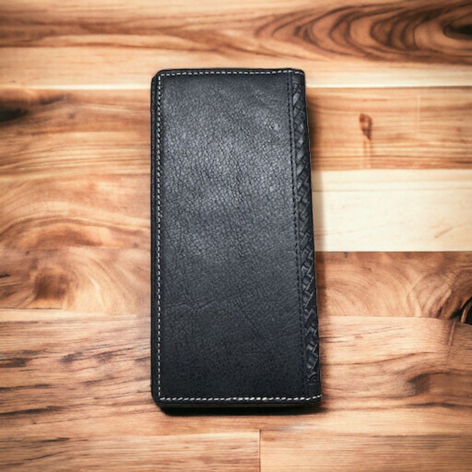 A black leather Lone Star Long Wallet Black W Cowhair Basketweave from Western Stakes with white stitching lies closed on a wooden surface with a mix of light and dark wood grains. The right edge of this cowboy accessory features a textured, braided pattern.