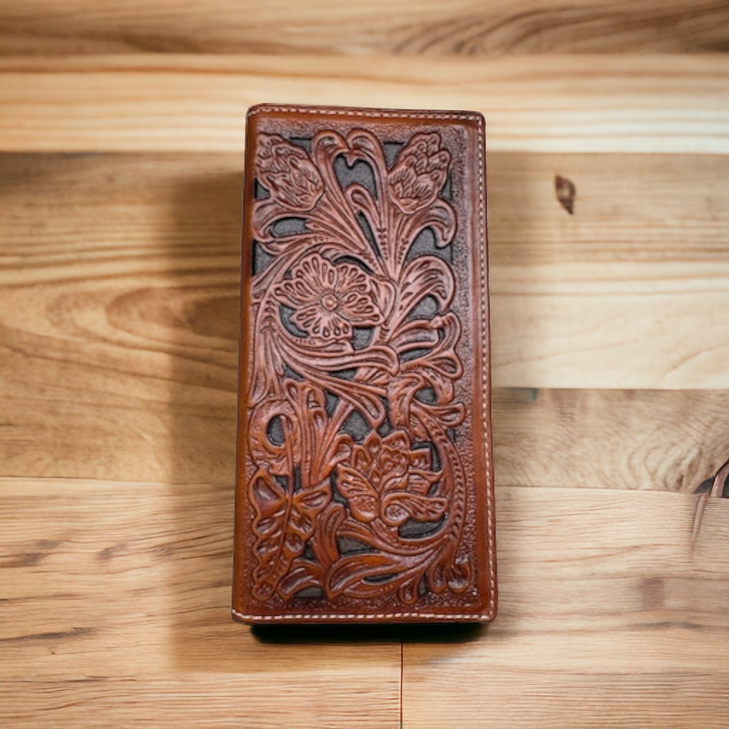 A detailed leather Floral Long Wallet Brown Dark Brown by Western Stakes rests on a wooden surface. The stylish man's accessory features intricate floral cutout designs embossed on its front, showcasing rich texture and craftsmanship. The wood beneath it has varied light and dark tones, adding contrast to the rustic appearance.