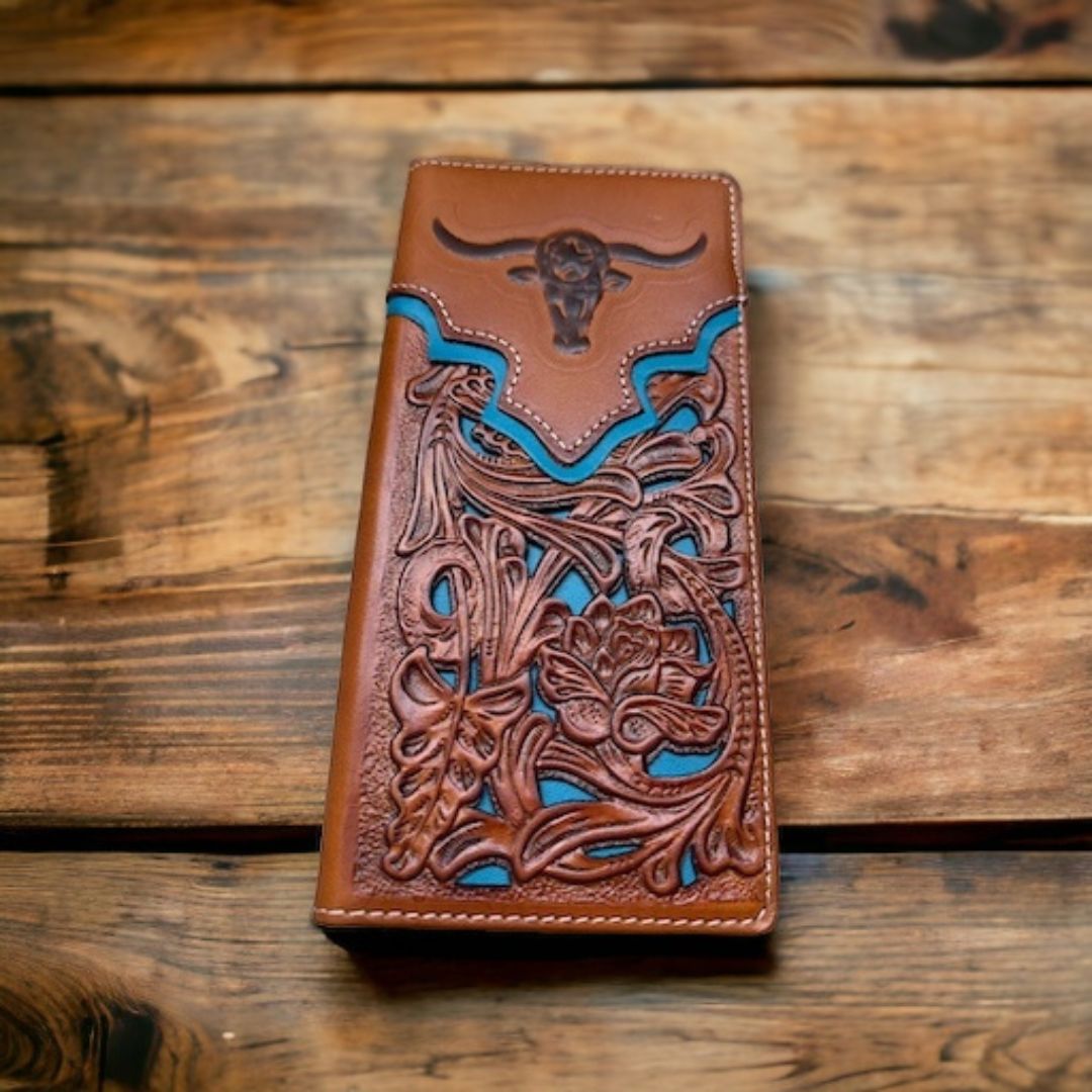 A Western Stakes Floral Long Wallet Longhorn Teal Accent made of high-quality leather, featuring intricate floral and Southwestern-style designs, including a longhorn skull motif, lies on a rustic wooden surface. The blue accents create a striking contrast with the brown leather, making it an ideal cowboy gift.