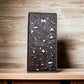 A dark brown leather wallet with intricate floral carvings and a checkbook style design stands upright on a wooden surface. The background has a blurred wood grain pattern, giving the image a rustic feel. This is the Floral Long Wallet Coffee White by Western Stakes.