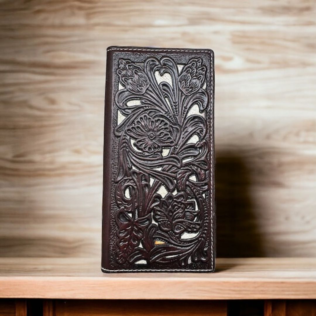 A dark brown leather wallet with intricate floral carvings and a checkbook style design stands upright on a wooden surface. The background has a blurred wood grain pattern, giving the image a rustic feel. This is the Floral Long Wallet Coffee White by Western Stakes.