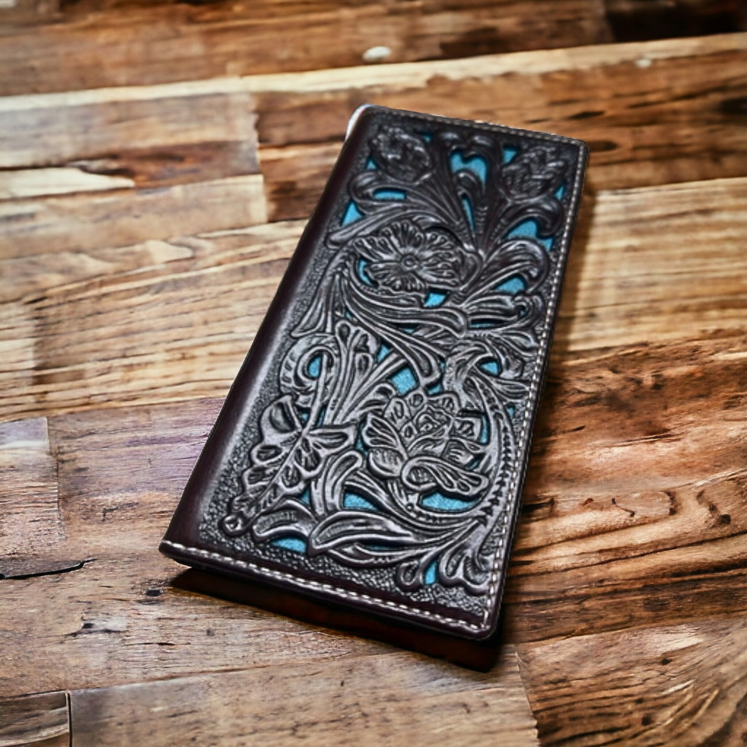 A finely detailed brown leather wallet with intricate floral patterns and hints of turquoise is placed on a wooden surface. The craftsmanship in the embossing highlights the beauty of the Floral Leather Long Wallet Coffee Teal design by Western Stakes.