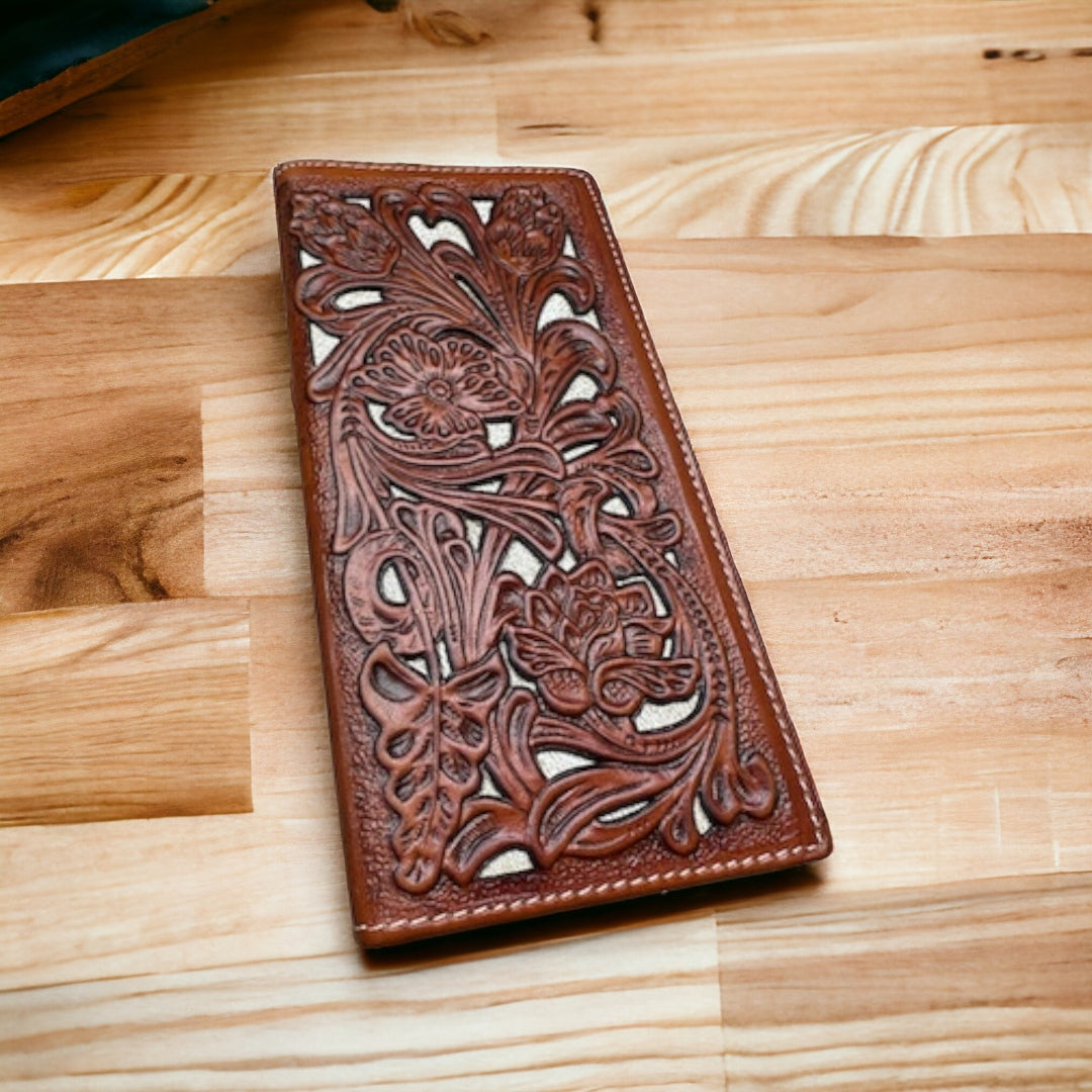 A Western Stakes Floral Leather Long Wallet Brown White with intricate floral designs embossed on its surface, resting on a light wooden table. The wallet's detailed pattern features leaves and flowers, creating a rich textured appearance reminiscent of a rodeo concho.