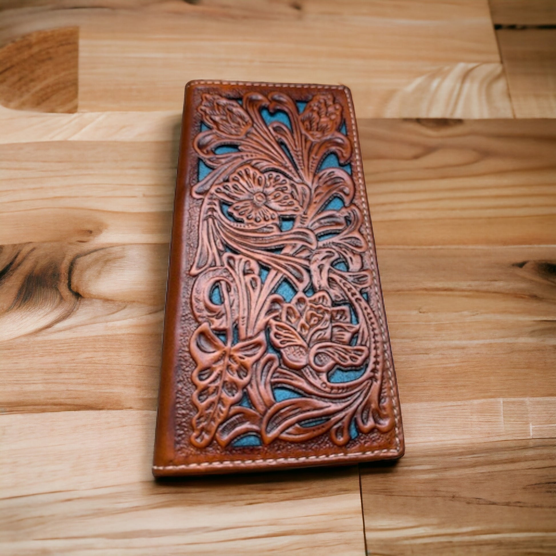 A Western Stakes Floral Leather Long Wallet Brown Teal featuring intricate floral and leaf designs. The design is carved into the leather, revealing a contrasting blue background. The wallet, resembling a floral cutout wallet, is placed on a wooden surface with various shades of wood grain visible.