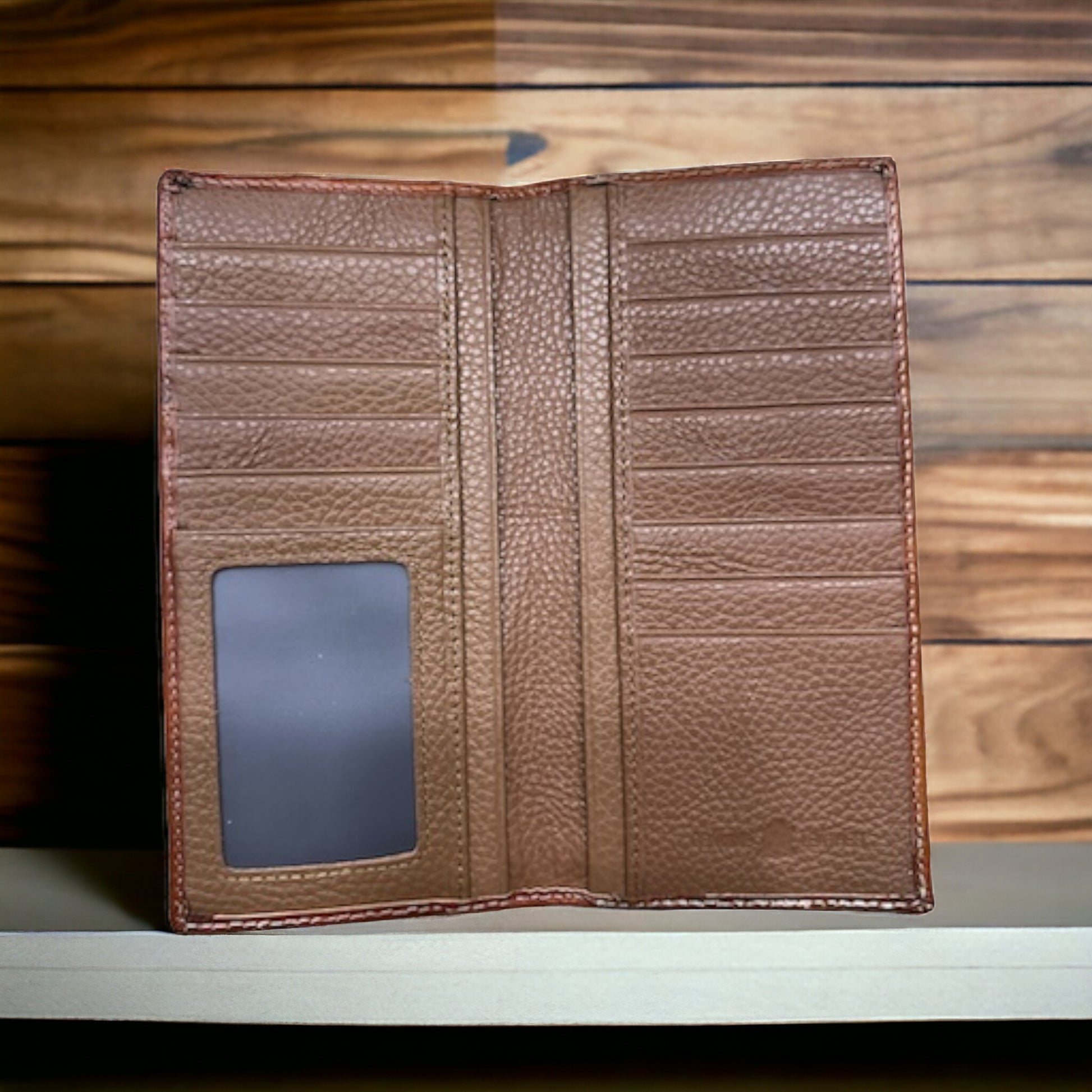 A Western Stakes Floral Leather Long Wallet Brown Teal is displayed on a wooden surface. The brown, open wallet features multiple card slots on each side and a clear ID slot on the bottom left. The fine craftsmanship shines against the wooden planks in various shades of brown.