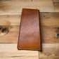 A Western Stakes Floral Leather Long Wallet Brown Teal with visible stitching along the edges lies closed on a wooden surface. The wallet appears worn, with some minor scuffs and creases, indicating frequent use. The wooden surface underneath has a mix of light and dark tones, adding texture to the image.