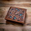 A Western Stakes Floral Leather Cowboy Wallet Brown & Teal with intricate floral cutout design and blue accents is placed on a wooden surface. The detailed design showcases craftsmanship and elegance, making it perfect for rodeo gifts.