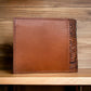 A brown leather book with intricate embossed designs on the spine stands upright on a wooden shelf, much like a Western Stakes Floral Leather Cowboy Wallet Brown & Teal. The rest of the cover is plain and unadorned. The background features horizontal wooden paneling, creating a warm, rustic ambiance.