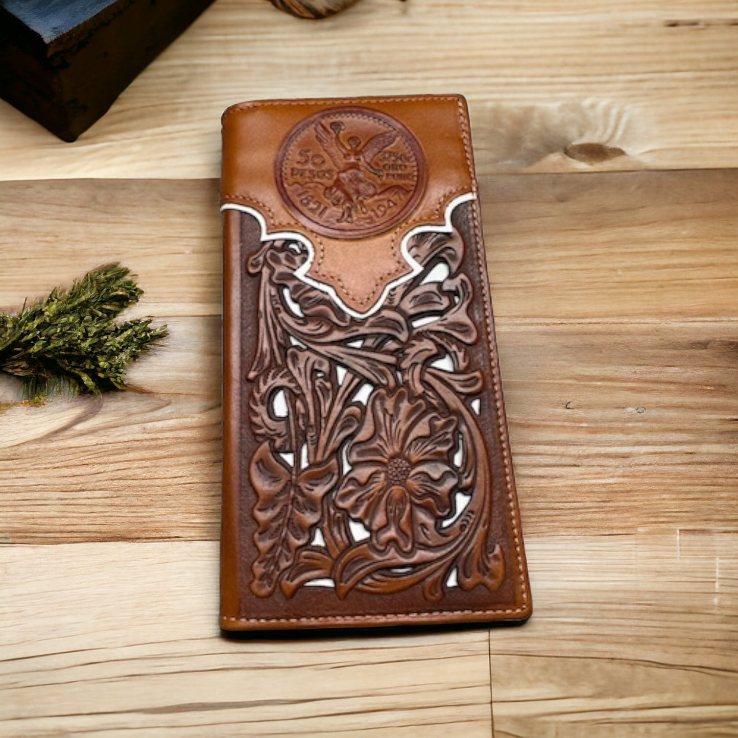 A hand-crafted Floral Long Wallet Brown White 50 Pesos from Western Stakes features intricate floral embossing and a circular Mexican 50 Peso design from Naciones Unidas Por Puebla 1979. The elegant wallet rests on a wooden surface next to a small bunch of dried flowers.