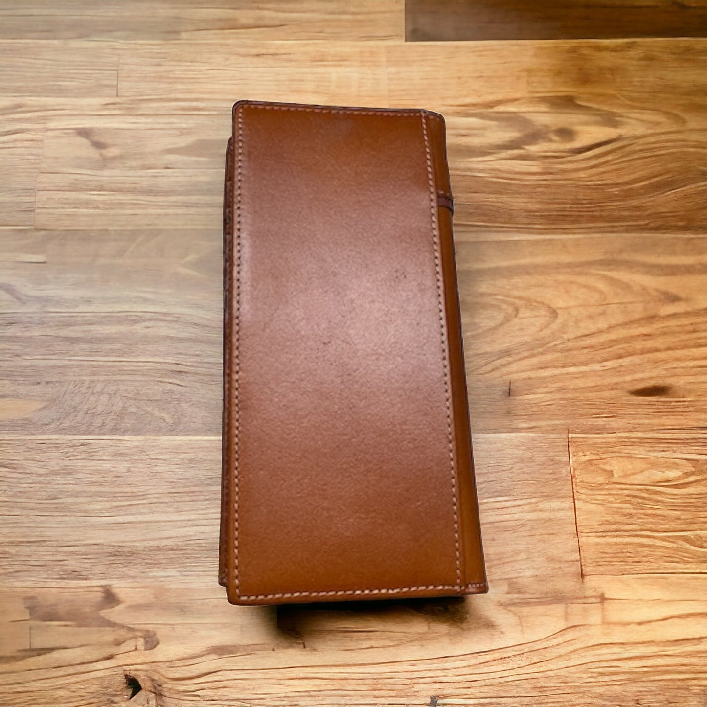 A Floral Long Wallet Brown White 50 Pesos by Western Stakes is placed closed on a wooden surface. The wallet, adorned with visible stitching around the edges and a smooth texture, features a subtle floral cutout design. The wooden surface has a varied grain pattern and a warm tone.