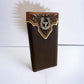 A sleek, rectangular Western Stakes Mens Leather Long Wallet Longhorn Horseshoe Coffee stands upright against a plain white background. It features an intricate metal emblem with a longhorn horseshoe concho and a bird, bordered with detailed scrollwork, and has a light brown and cream decorative trim on the top edge.