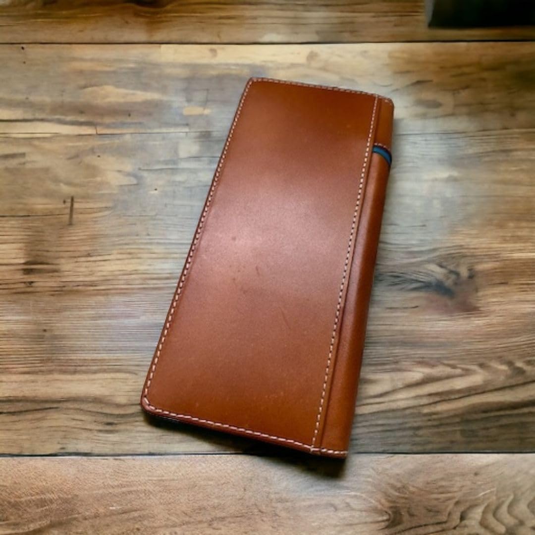 A high-quality Floral Long Wallet Longhorn Teal Accent from Western Stakes with a small elastic loop holding a pen is placed on a rustic wooden surface with natural grain patterns. The wallet has clean, white stitching around its edges, giving it a simple yet elegant look—an ideal cowboy gift for those who appreciate craftsmanship.