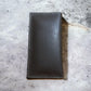 A stylish, high-end brown leather wallet with visible white stitching along the edges is placed on a slightly weathered, speckled concrete surface. The wallet appears closed and is oriented vertically in the center of the image, reminiscent of a Western Stakes Horseshoe Long Wallet Coffee Red Trim W Cowhair.