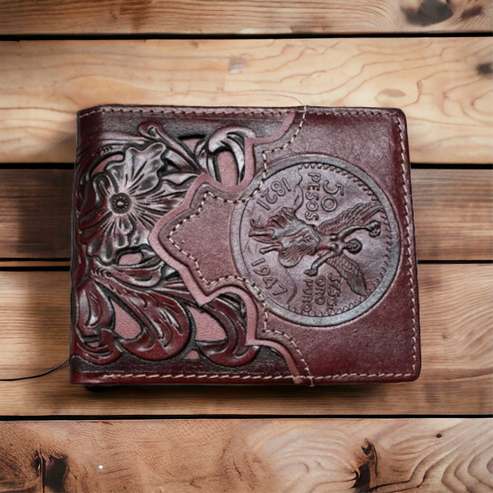 A handcrafted Western Stakes Floral Leather Cowboy Wallet Coffee 50 Pesos with a floral cutout design and a large embossed coin-like detail in the center, featuring the years 1911 and 1947. The leather wallet sits on a wooden surface marked with horizontal planks.