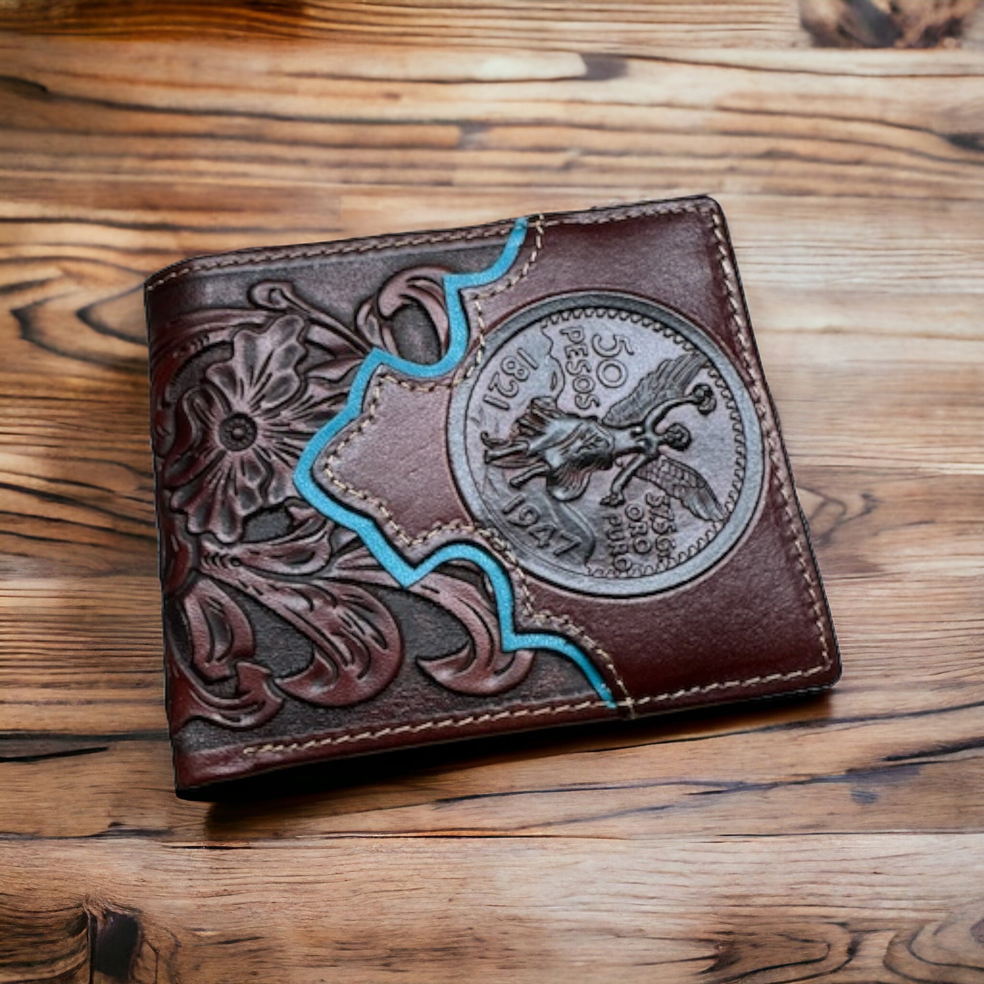 A Floral Leather Cowboy Wallet Coffee Teal 50 Pesos by Western Stakes rests on a wooden surface. The brown leather features an intricate floral cutout design and an embossed, circular emblem with the number "50" and "1967" on it, as well as a depiction of a figure in action and blue accents.