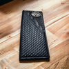 A detailed black leather Bull Rider Long Wallet W Cowhair Basketweave from Western Stakes with a woven texture and an ornate silver rodeo concho emblem in the top corner lies on a polished wooden surface. The wallet has intricate stitching and design elements, giving it a sophisticated and handcrafted appearance.