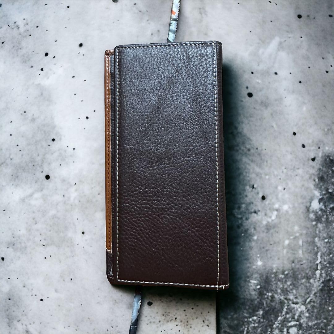 A closed, dark brown **Mens Long Wallet Coffee Longhorn Concho** from **Western Stakes** with visible stitching sits on a speckled concrete surface. The wallet's rugged texture exudes western charm, and there is a contrasting lighter brown leather strip on one side.