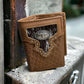 A Western Stakes Mens Brown Tri Fold Leather Wallet Longhorn Concho is propped against a metal surface. The wallet boasts intricate stitching, a vertically woven leather pattern, and cow hair leather details, showcasing its well-crafted and rugged design.