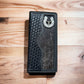 A Longhorn Long Wallet Black With Cowhair Basketweave by Western Stakes features a detailed silver horseshoe ornament at the top and is placed on a wooden surface.