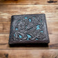 A **Western Stakes Floral Leather Cowboy Wallet Brown & Teal** is placed on a wooden surface. The embossed design includes detailed flowers and leaves, accented by a blue background behind the floral pattern.