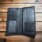 An open black leather wallet is displayed on a wooden surface. The wallet features card slots on the left side, a transparent ID slot at the bottom left, and two large pockets on the right side. The brand name and logo are embossed at the bottom right, showcasing the distinctive Longhorn Long Wallet Black With Cowhair Basketweave design by Western Stakes.