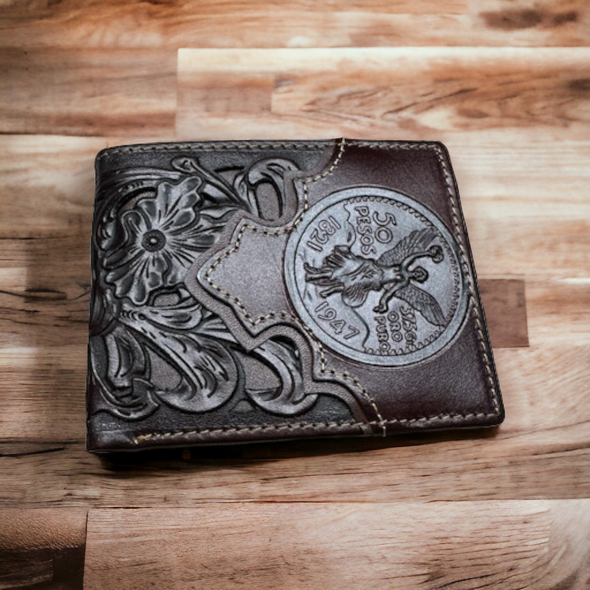 A Floral Leather Cowboy Wallet Coffee Brown 50 Pesos from Western Stakes with intricate floral engravings and a prominent Mexican 50 Peso coin design from 1947 embossed on its cover. The men's Western wallet is set on a wooden surface with varying shades of light and dark wood grain.