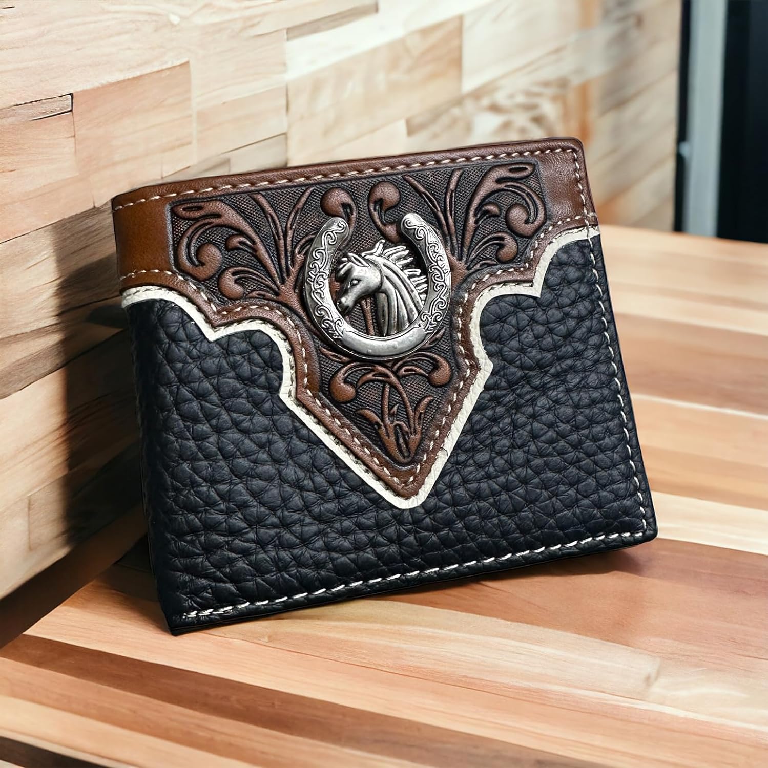 A Western Stakes Mens Leather Wallet Horse & Horseshoe Soft Black Leather Bi Fold with a black and brown leather finish, featuring intricate embossed designs and an engraved horse head emblem in the center. The top part showcases detailed craftsmanship with a horse motif, while the lower part has a textured black leather finish. The Western Stakes mens leather wallet rests on a wooden surface.