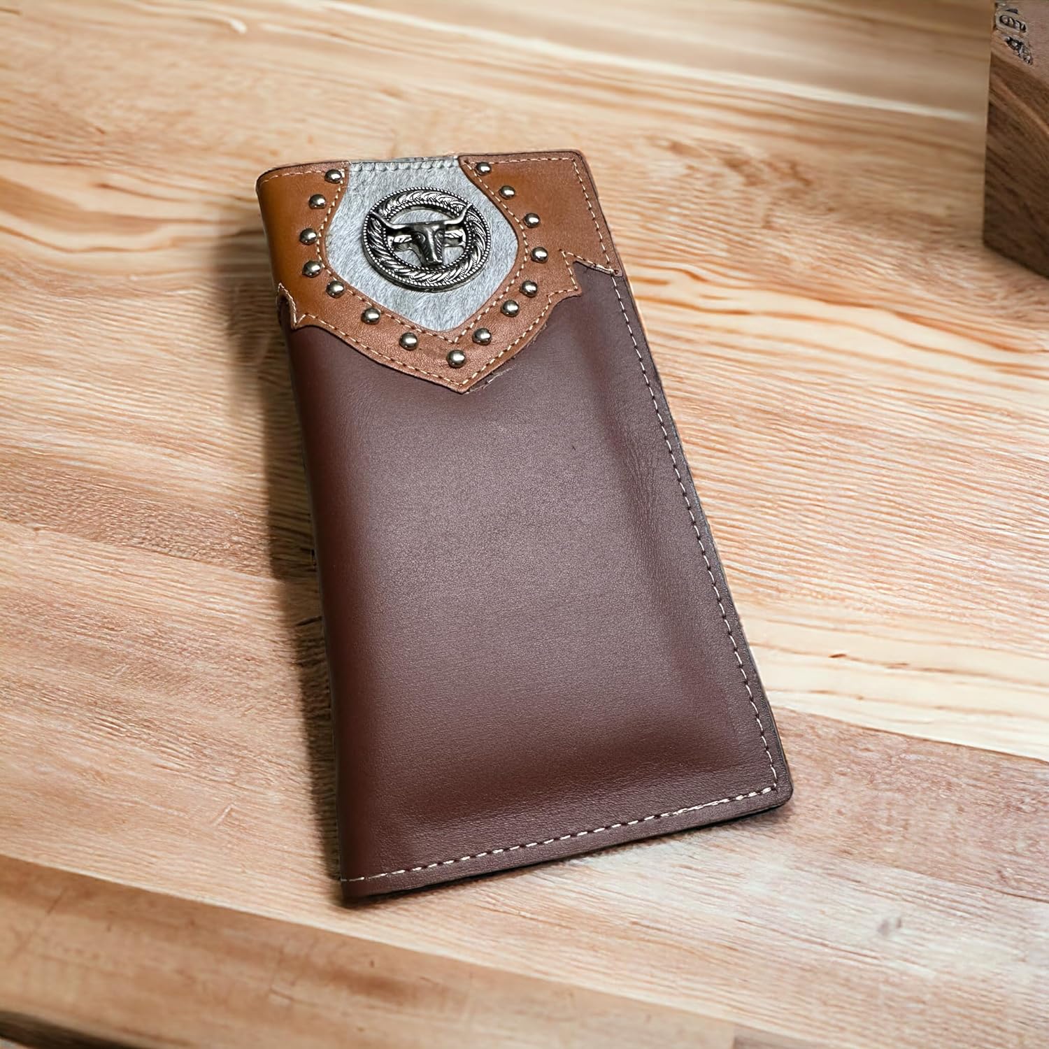 A brown leather wallet lies on a wooden surface. The Western Stakes Longhorn Long Wallet Brown With Cowhair & Concho features a decorative silver embellishment with a bull's head design and stud accents at the top. This western accessory rests against a background showing a light-colored wood grain pattern.