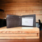 An open black Mens Leather Wallet Horseshoe Longhorn Coffee Bi Fold by Western Stakes displaying a card with the text "NO:211 Colorcoffee" rests atop a wooden table, against a light-colored, textured wood wall. This stylish bi-fold design adds a rugged touch to any ensemble.