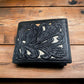 A black leather checkbook-style wallet with an intricate floral cutout design is placed on a wooden surface. The pattern is detailed with visible textures, adding depth to the aesthetic. This handcrafted piece exudes stylish uniqueness and cowboy charm. The Floral Leather Cowboy Wallet Brown & Teal by Western Stakes adds a captivating rustic elegance to any ensemble.