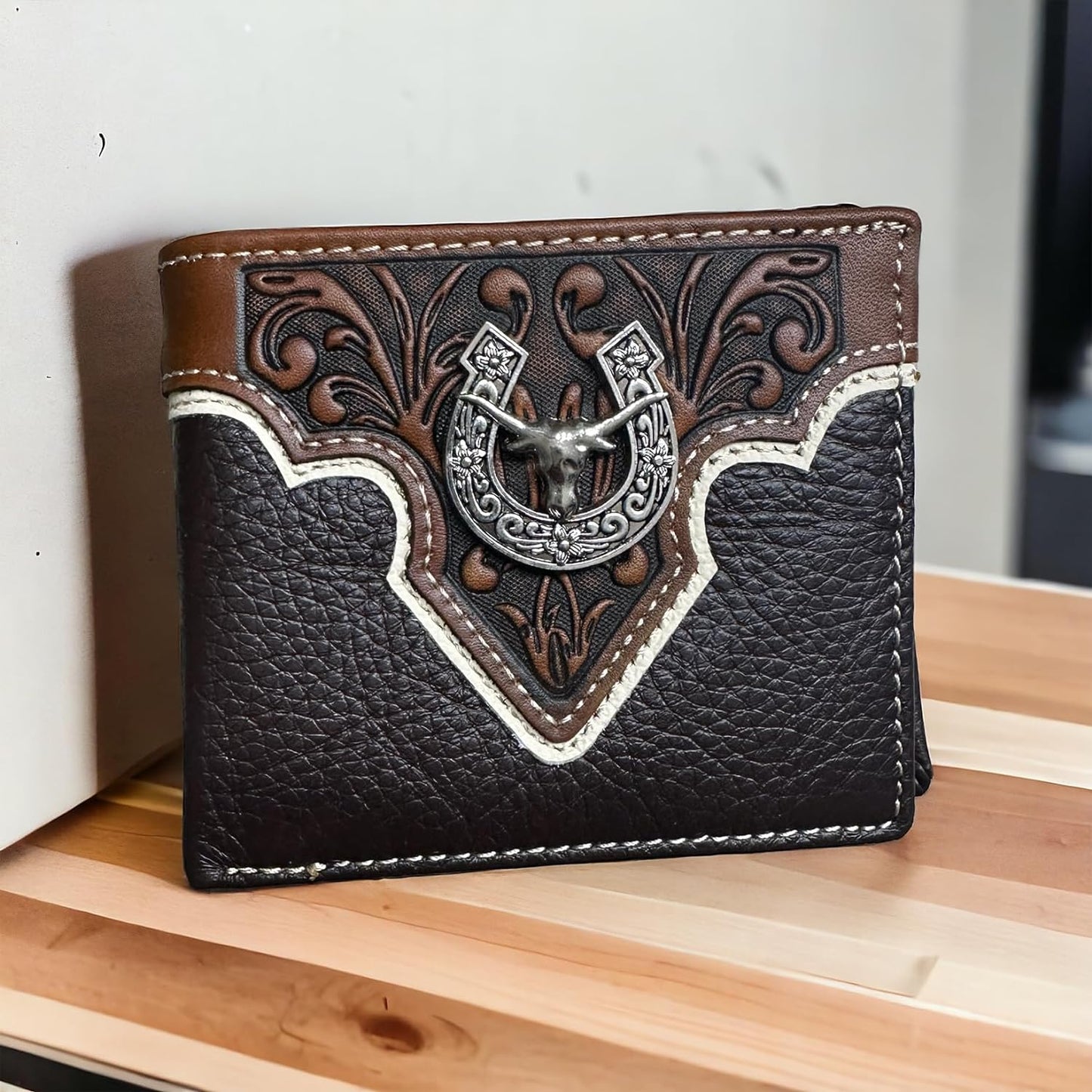 A stylish Mens Leather Wallet Horseshoe Longhorn Coffee Bi Fold from Western Stakes features an intricate embossed design with a horseshoe emblem on the front. This western cowboy wallet has decorative stitching in contrasting white, showcasing a blend of dark and light brown shades. The bi-fold design is displayed standing on a wooden surface.