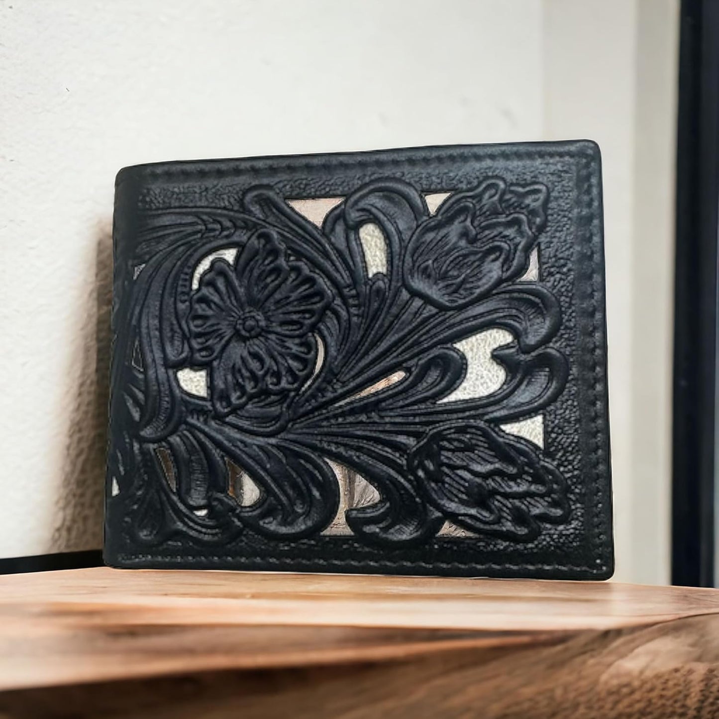 A handcrafted Western Stakes Floral Leather Cowboy Wallet Brown & Teal with a decorative floral cutout design sits upright on a wooden surface against a plain, light-colored wall. The intricate floral pattern is embossed and visually prominent on the wallet's exterior.