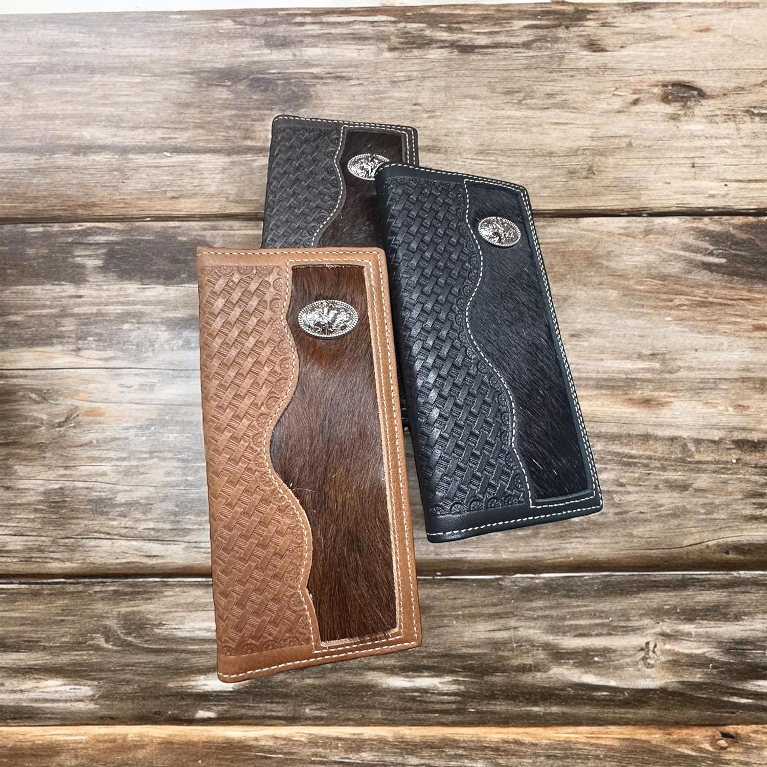 Three intricately designed Bull Rider Long Wallet W Cowhair Basketweave by Western Stakes are displayed on a wooden surface. The wallets, perfect as a cowboy accessory, feature a combination of leather and woven textures in black and brown shades. They have silver metallic emblems at the top right corner. The background is a rustic wooden pattern.