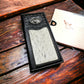 A fashionable Western Stakes Rodeo Long Wallet Black With Cowhair & Bull Rider Concho with intricate stitching and a light-colored cowhide patch on its front is placed on a wooden surface. A small decorative metal emblem is embedded in the upper part of the checkbook style wallet. Nearby, a partially visible notebook with a butterfly design lies on the table.