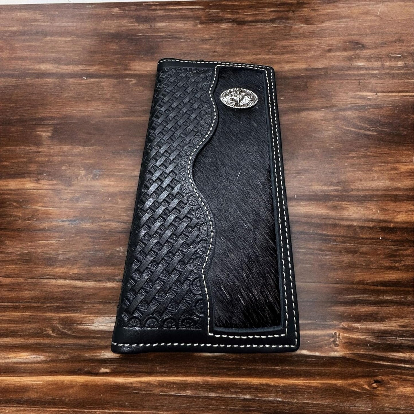 The Western Stakes Bull Rider Long Wallet W Cowhair Basketweave is a stylish black leather wallet with an intricate woven design and a smooth, hair-on-hide section. It features white stitching around the edges and a silver oval concho embellishment with a horse design on the upper right corner. This cowboy accessory rests on a wooden surface.