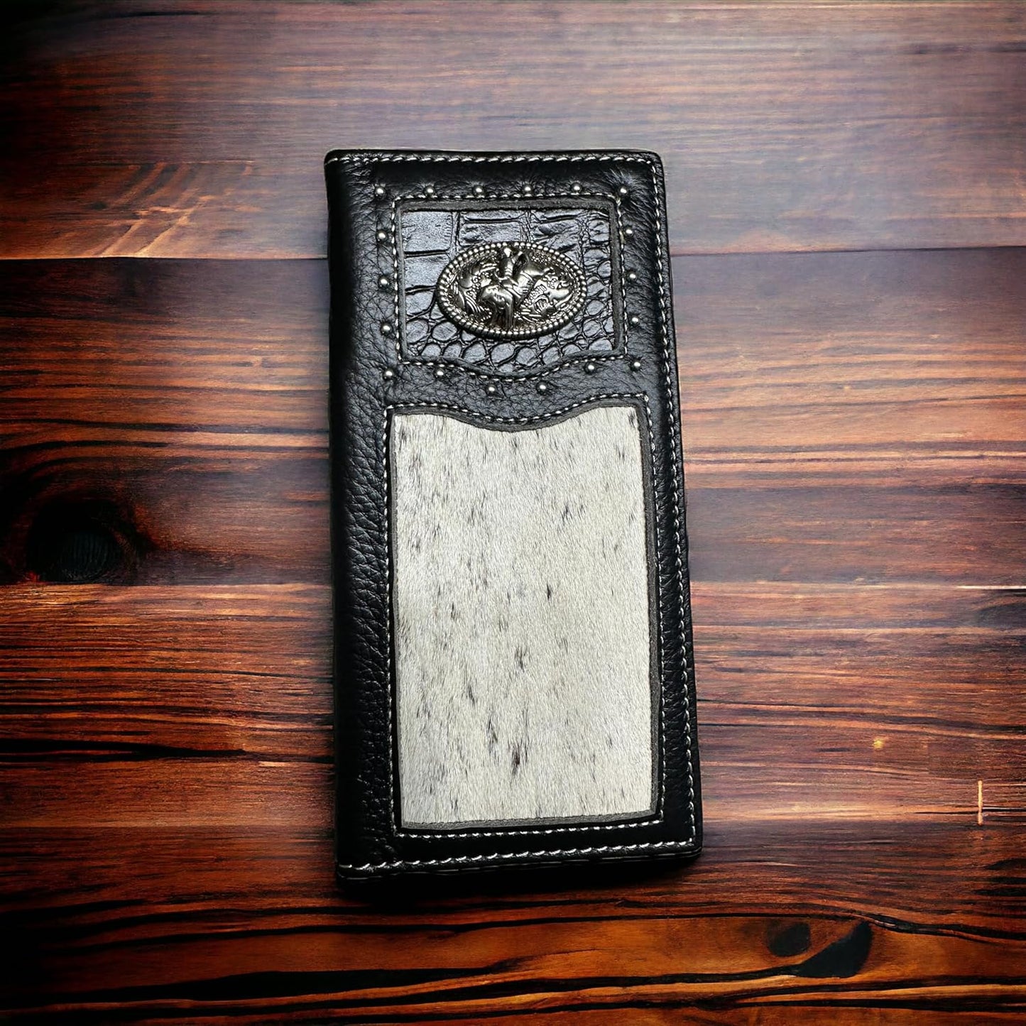 A rectangular black leather wallet lies on a wooden surface. This fashionable Western Stakes Rodeo Long Wallet Black With Cowhair & Bull Rider Concho features an intricate embossed design at the top and a large, light grey textured patch in the center. The wooden background has a rich, warm grain pattern.